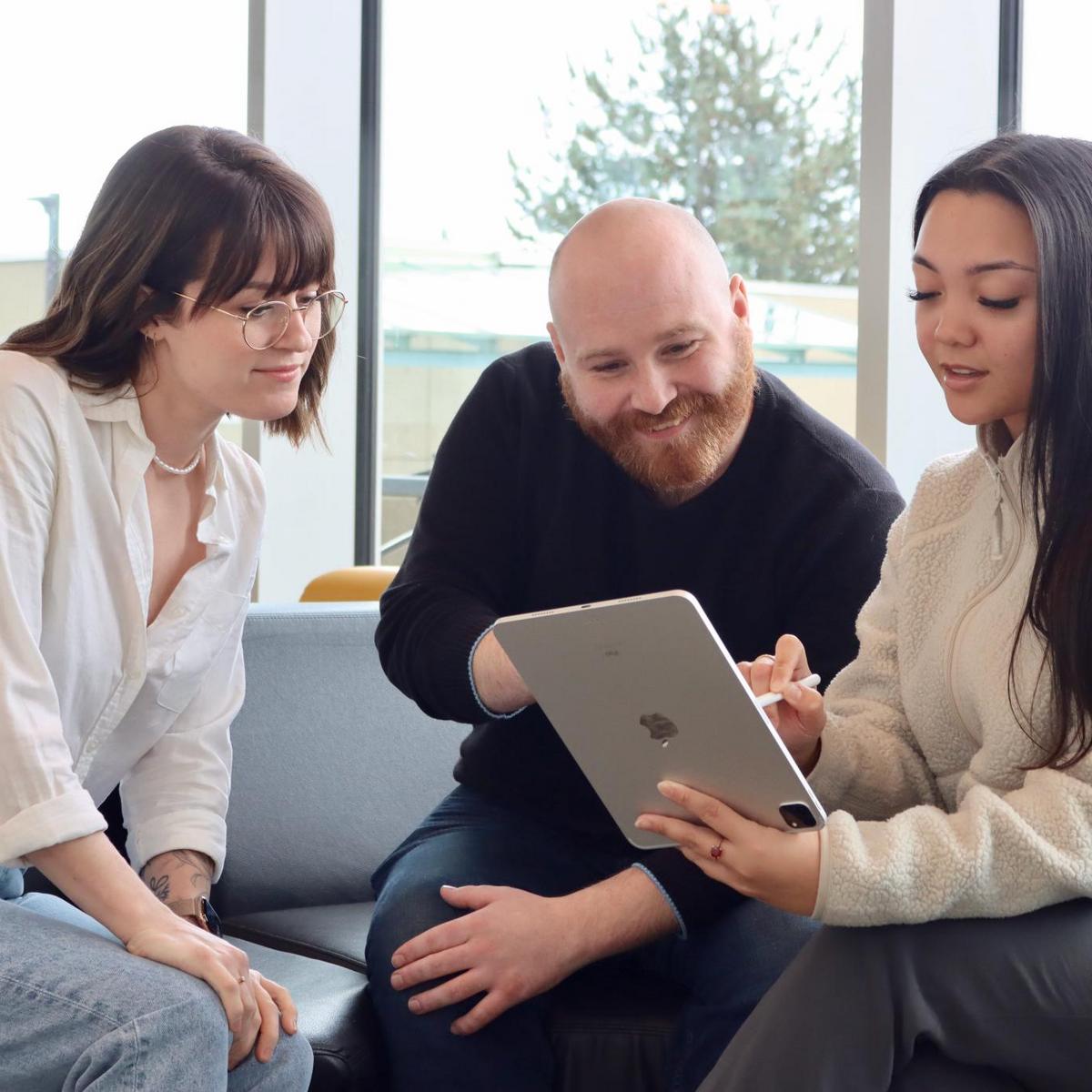 A woman, a man and another woman sitting on a chesterfield looking at a laptop together.