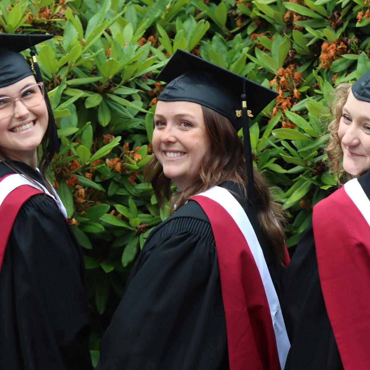 Three students wearing caps and gowns in front of a bush