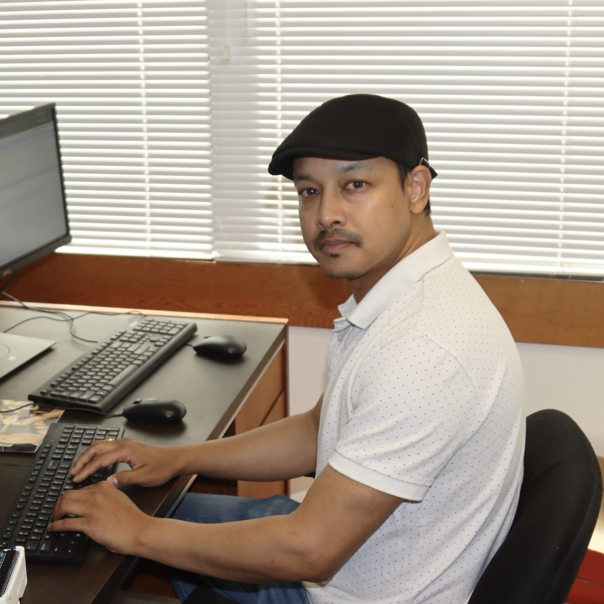Ajay Shrestha sitting in his office, looking at the camera and in front of a keyboard and three computer screens