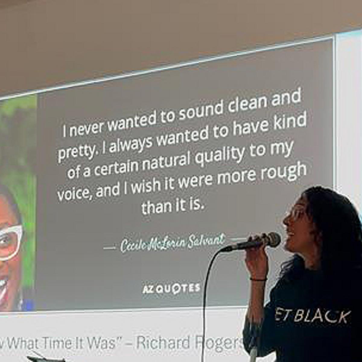 A Black woman speaks into a microphone with a PowerPoint slide behind her
