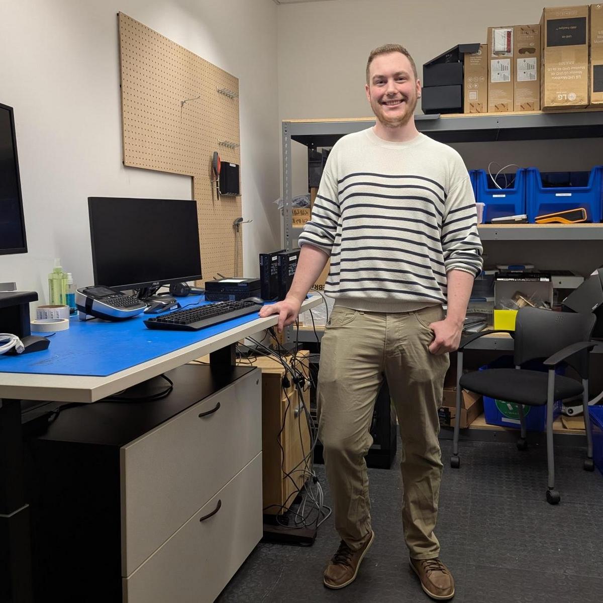 A man standing beside a desk with a computer and a monitor on it in a room with shelves and supplies on them