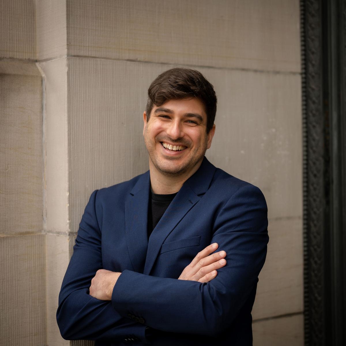 Michael MacKenzie standing against a wall and smiling at the camera.