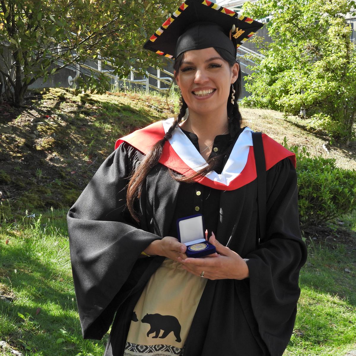 VIU Graduate, wearing convocation robes, Olivia Thomas is holding a medal while standing outside in front of a garden 
