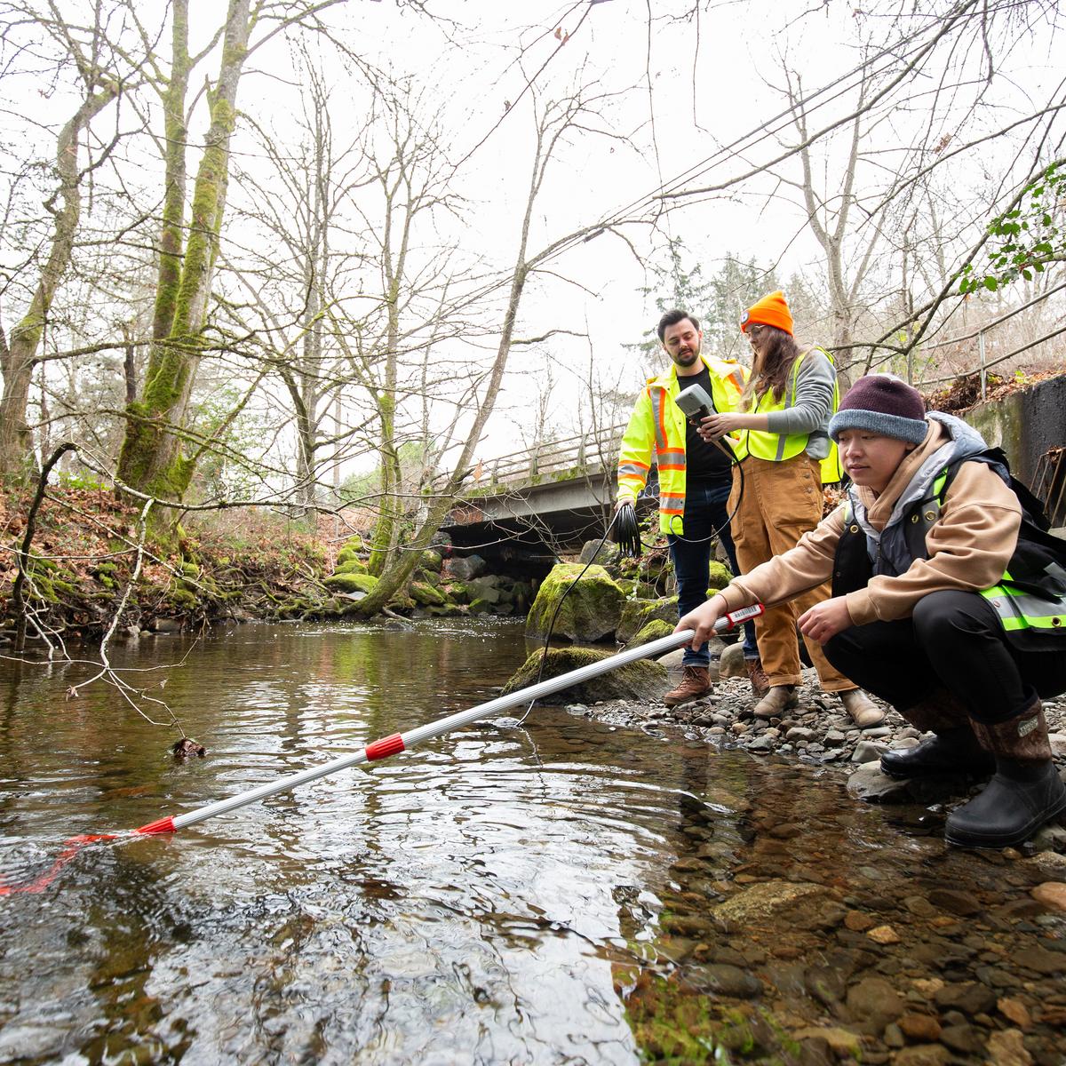 Three people next to a stream, one holds a sampling pole while the others look on