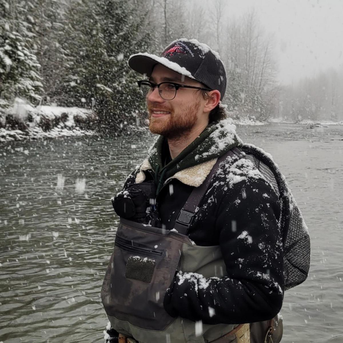 A young man clad in hipwaders is standing in a river with snowflakes falling around him