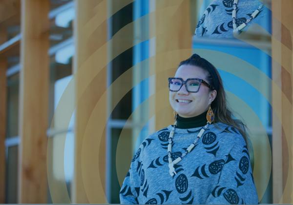 A smiling young woman wearing  Indigenous-themed top and jewelry