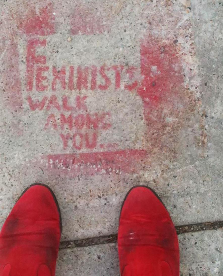 A pair of red shoes stand on a light grey concrete sidewalk that is spray painted with the phrase "Feminists Walk Among You." 