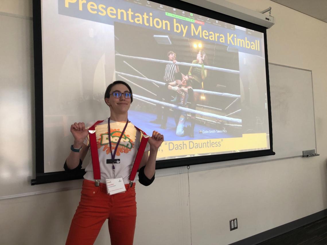 Meara Kimball stands in front of a projector screen in a wrestling costumne for a presentation on prior learning 