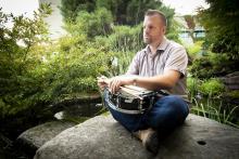 VIU Music Department Instructor Hans Verhoeven sits on a rock near a pond holding a drum.