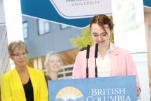 VIU student Mallory Woods stands at a podium wearing a pink blazer. VIU banners stand behind them.