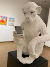 An ape made of white plaster and clay holds a cell phone, which it stares at intensely