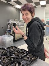 Melanie P. Loureiro stands in front of several bins of mussels and holds onel in her left hand.