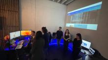 Students gather around three screens on a desk with a projection of an image on the wall behind them.