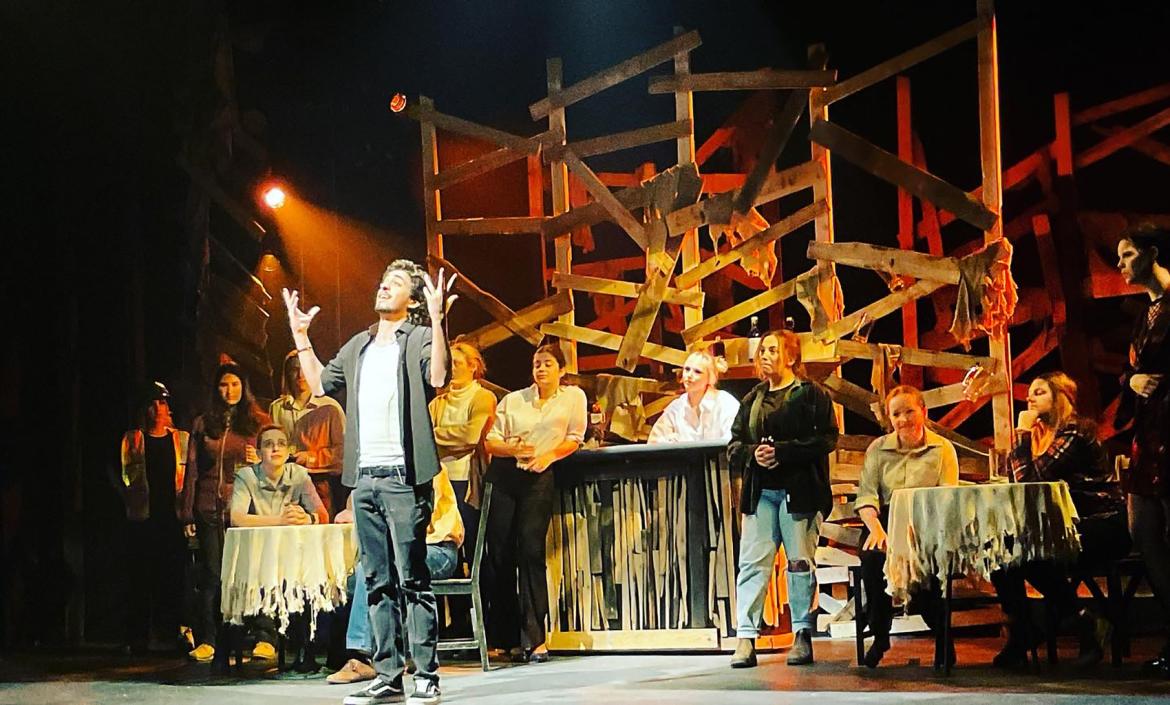 An actor at centre stage has their hands thrown up in the air while they look off in the distance. Numerous cast mates stand behind them in various poses.