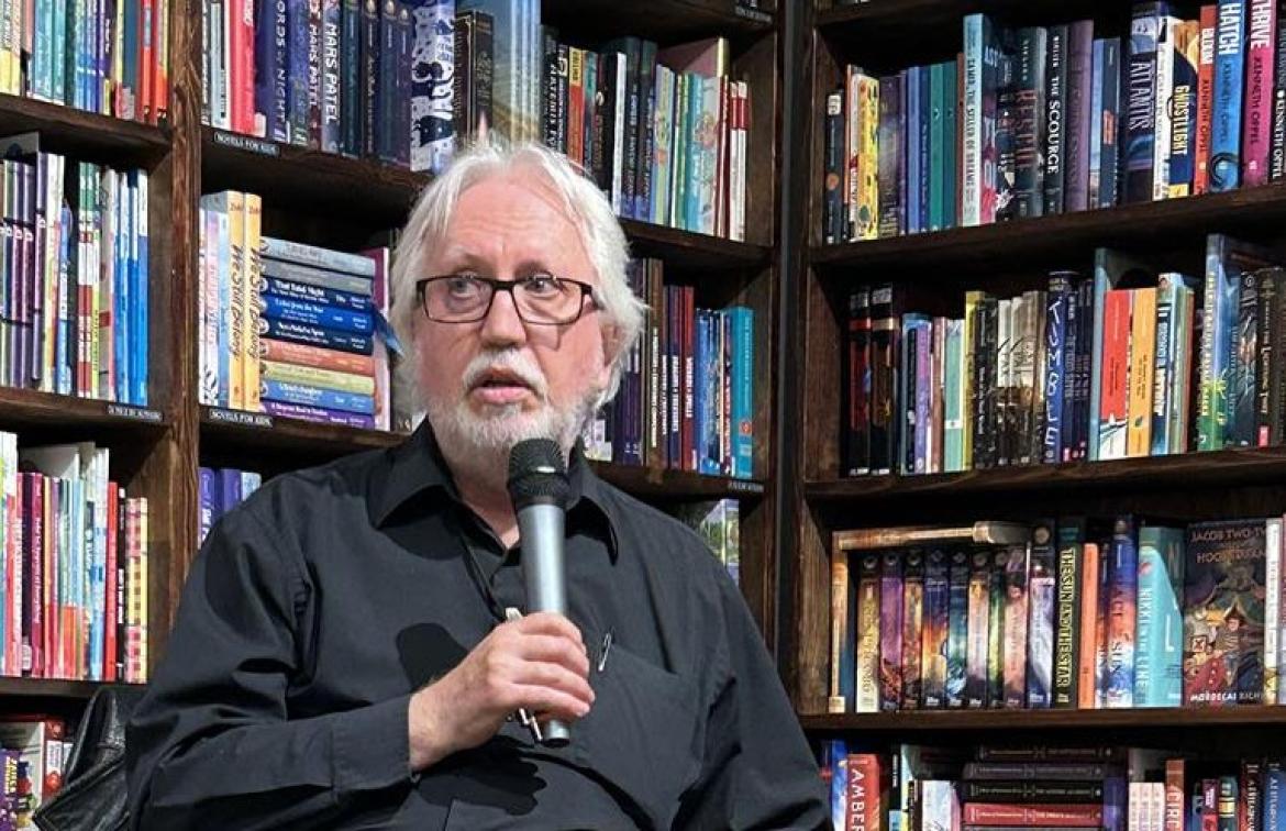 Perry Bulwer speaks into a microphone with a shelf of books behind him