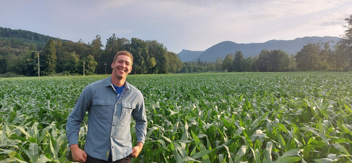 Douglas Groenendijk grins while standing in a vast green field with a mountain range behind him.