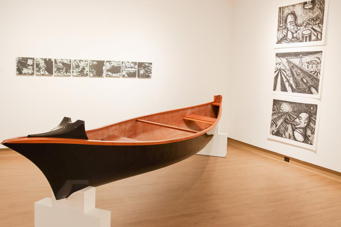 A canoe sits on a stand with drawings hanging on the gallery wall's behind it.