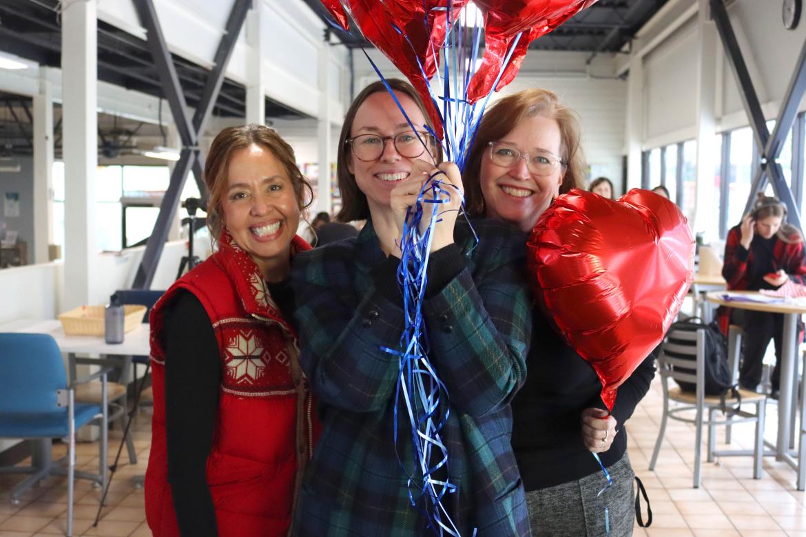 Three women smile, one holding a bunch of balloons, another holding a heart balloon near her face