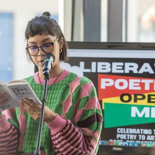 A woman wearing glasses and a pink and green sweater reads from a poetry book at a microphone