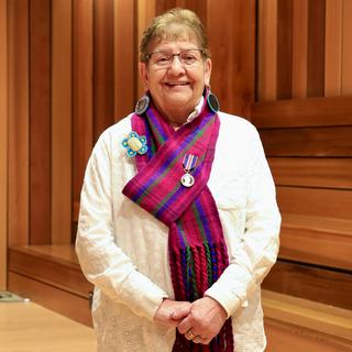 Métis Elder-in-Residence Stella Johnson standing in a cedar panelled room wearing a red and purple tartanMétis scarf with a King Charles III Coronation Medal