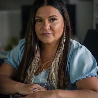 Brandi is wearing a blue shirt and sits with her arms on a table in front of her