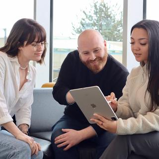 A woman, a man and another woman sitting on a chesterfield looking at a laptop together.