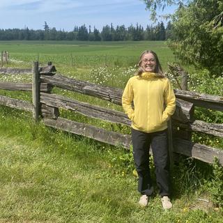 A woman stands next to a fence and a green field with her hands in her pockets