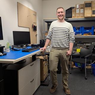 A man standing beside a desk with a computer and a monitor on it in a room with shelves and supplies on them
