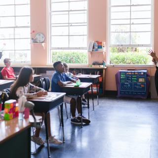 Students sit at desks in a class listening to a teacher