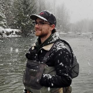 A young man clad in hipwaders is standing in a river with snowflakes falling around him