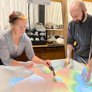 Gerri McEwen and Devin Ayotte dig through sand that is lighted blue, green, yellow and red.