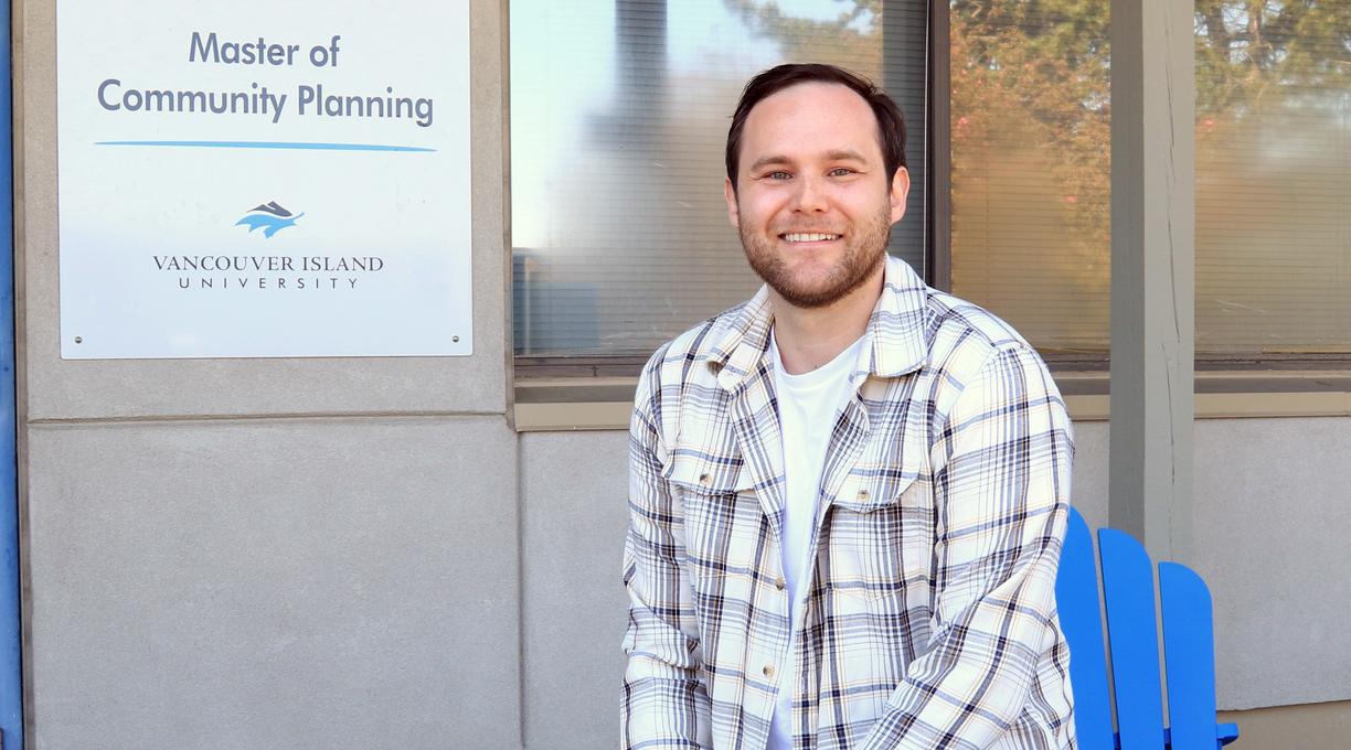 A man wearing a beige and white plaid jacket over a white t-shirt is sitting on the arm of blue Adirondack chair in front of a door and window to a building