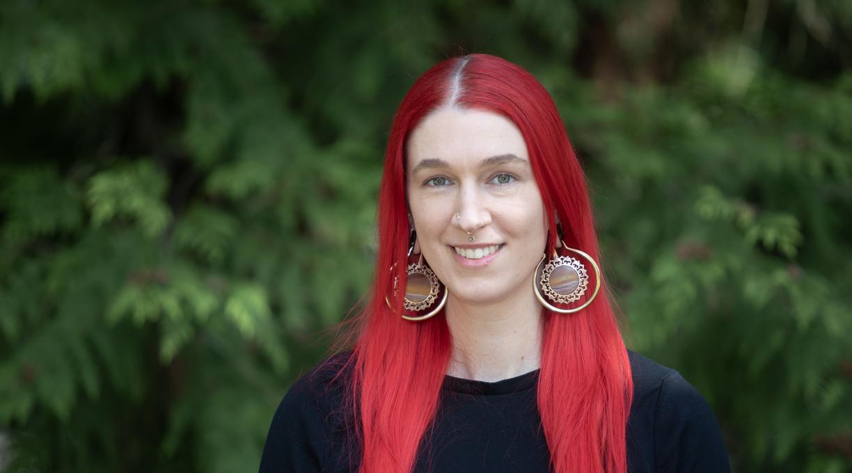 A young woman with bright red, long hair is smiling with tree leaves in the background