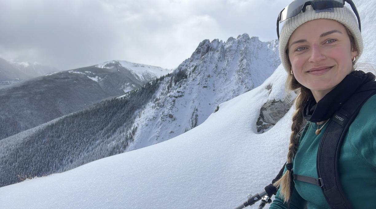 Andrea wearing ski gear smiles with a snowy mountainscape behind her