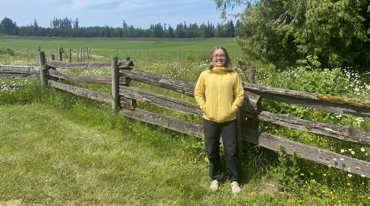 A woman stands next to a fence and a green field with her hands in her pockets
