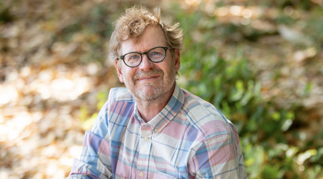 Erik Krogh sitting on the ground outside on a sunny day and smiling at the camera.