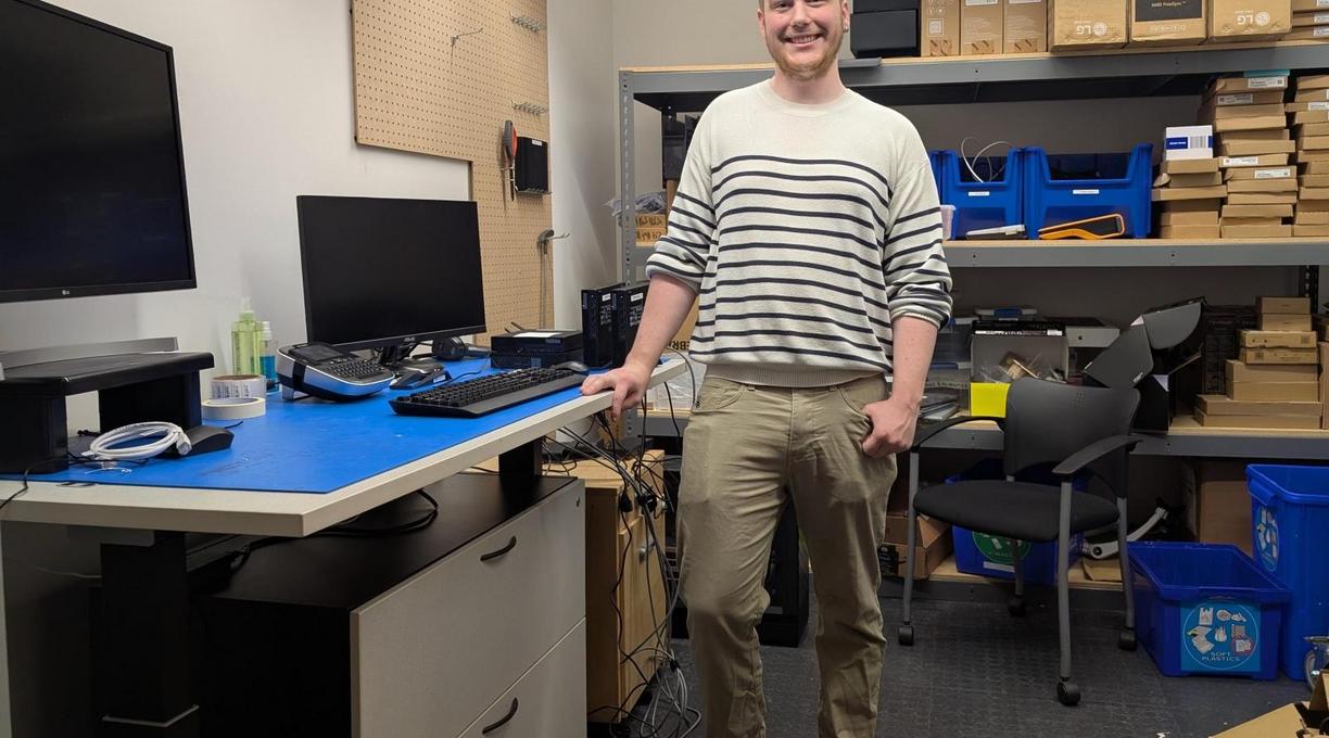 A man standing beside a desk with a computer and a monitor on it in a room with shelves and supplies on them