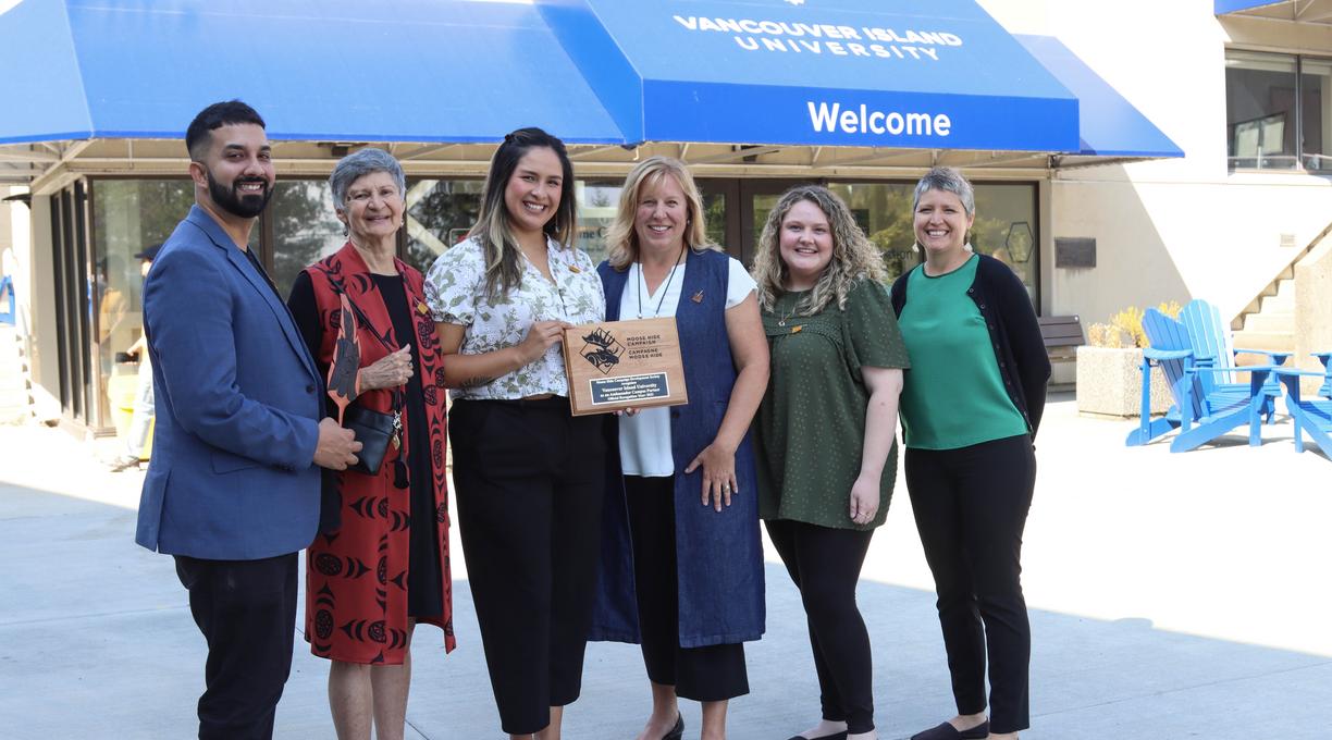 A group stands in front of the welcome centre holding a plaque