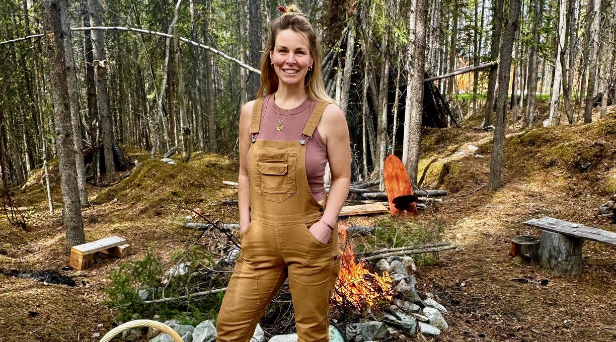 A woman wearing tan overalls is standing in a clearing in the forest in front of a campfire. 