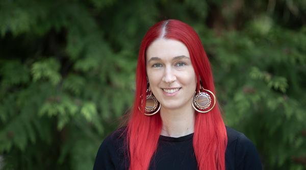 A young woman with bright red, long hair is smiling with tree leaves in the background