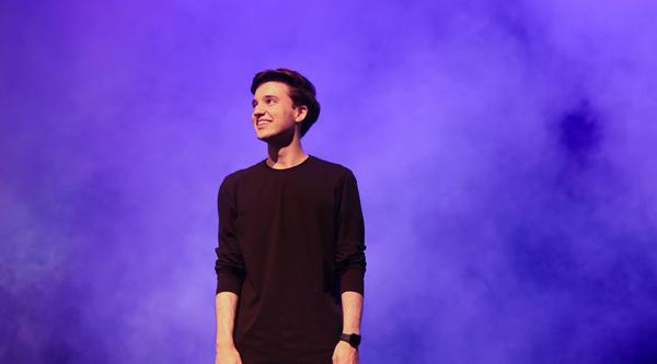 Alex Bhargava stands on the Malaspina Theatre stage looking off to the left. He wears all black and blue and purple smoke swirls around him.