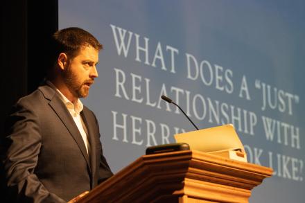 Man speaks at podium with text on a screen that reads: What does a just relationship with herring look like? 