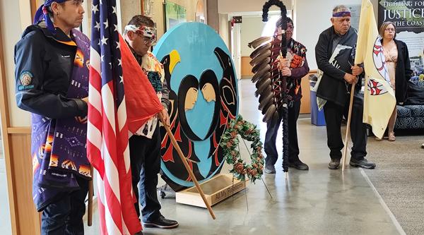 Indigenous community members wearing blankets stand solemnly next to cenotaph next to 