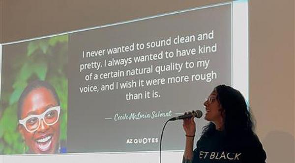 A Black woman speaks into a microphone with a PowerPoint slide behind her