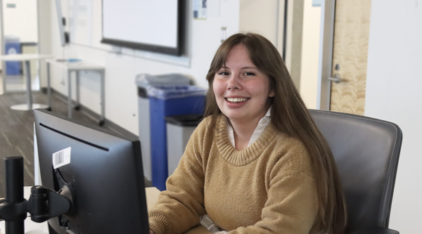 Kaylah Vassallo sittiing at a desk in front of a computer monitor and smiling at the screen.