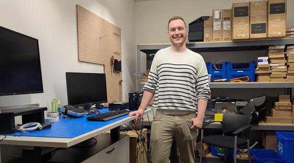 A man standing beside a desk with a computer and a monitor on it in a room with shelves and supplies on them