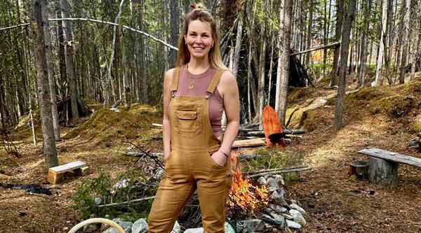 A woman wearing tan overalls is standing in a clearing in the forest in front of a campfire. 