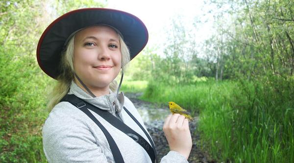 Chelsey Watts stands amongst bushes and trees holding a bird.