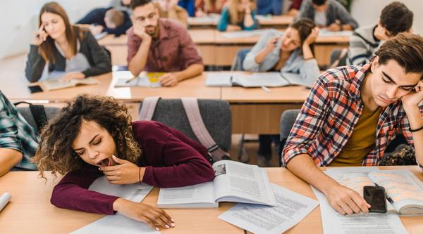 a student on the left yawns while the student next to her has his hand on his face while in a classroom with lots of students in the background