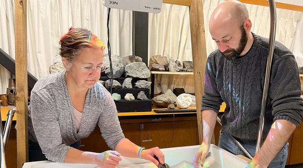 Gerri McEwen and Devin Ayotte dig through sand that is lighted blue, green, yellow and red.